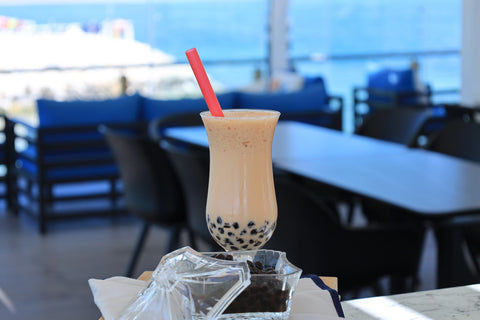 Chilled boba tea with tapioca balls and straw, set against a scenic seaside view in a modern café.