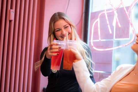 Two women smiling while toasting bubble tea in a vibrant indoor setting.