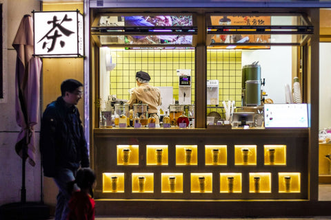 A man and child passing by a vibrant bubble tea shop at night in Nanjing, China.