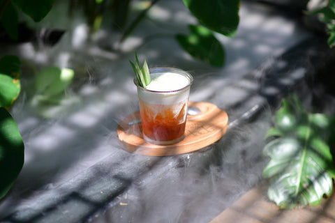 A glass of iced tea with foam and garnish in a misty garden setting with lush greenery.