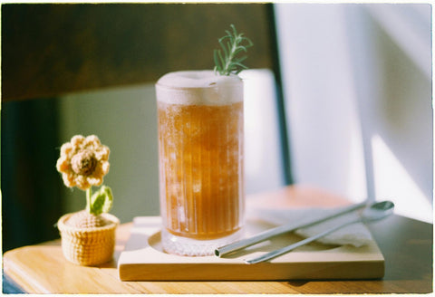 Glass of iced tea with foam and rosemary garnish on wooden table, accompanied by a cute fabric flower pot.