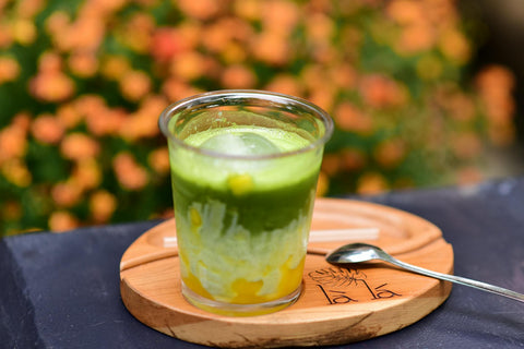 A vibrant matcha and mango iced drink set outdoors against blooming flowers, on a wooden tray.