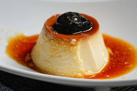 Creamy flan dessert topped with caramel sauce and a prune on a white plate.