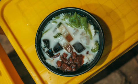 A vibrant Asian dessert in a black bowl showcasing colorful jellies and beans, set on a yellow table.