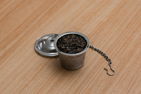 A stainless steel tea infuser containing loose leaf tea on a wooden surface.