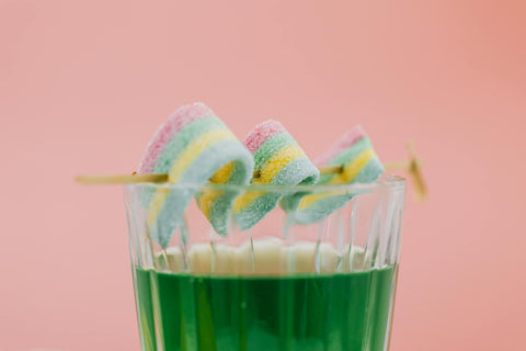 Colorful candy skewers dipped in green beverage against pink backdrop.