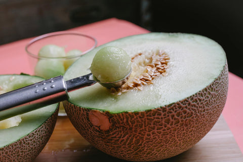 Half of fragrant melon with seeds placed on cutting board with scoop of flesh on pink table on blurred background