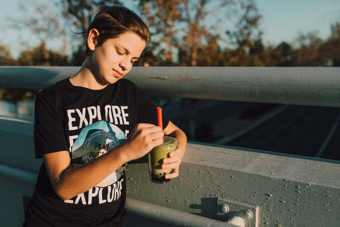 A teenager enjoys a boba milk tea on a sunny day, leaning on an outdoor railing.