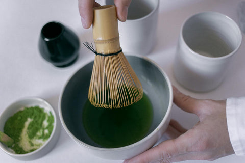 Close-up of hands using a bamboo whisk to prepare matcha tea in a ceramic bowl.