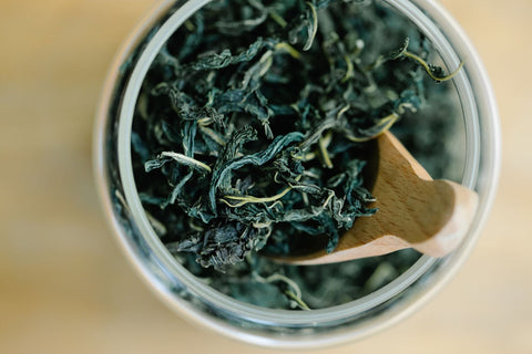 Top view of dried green tea leaves in a glass jar with a wooden spoon.