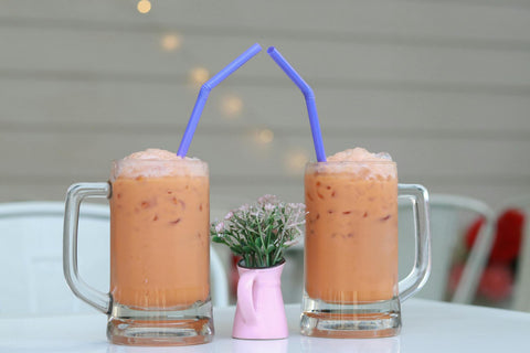 Two mugs of iced Thai milk tea with straws beside a small flower vase.